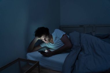Addicted Man sohbet ve yatakta gece geç saatlere kadar akıllı telefon ile Internette sörf. Sıkılmış, uykusuz ve kasvetli ışık ile karanlık odada yorgun. Uykusuzluk ve mobil bağımlılık kavramı.