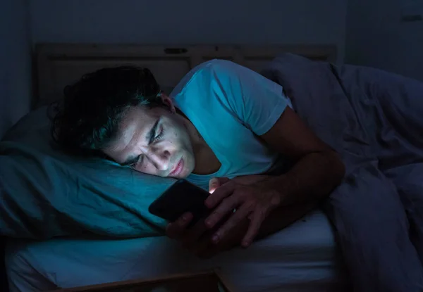 Addicted man chatting and surfing on the Internet with smart phone late at night in bed. Bored, sleepless and tired in dark room with moody light. In insomnia and mobile addiction concept.