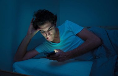 Addicted Man sohbet ve yatakta gece geç saatlere kadar akıllı telefon ile Internette sörf. Sıkılmış, uykusuz ve kasvetli ışık ile karanlık odada yorgun. Uykusuzluk ve mobil bağımlılık kavramı.