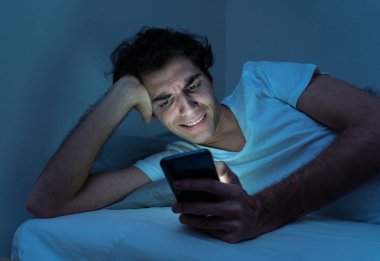Addicted Man sohbet ve yatakta gece geç saatlere kadar akıllı telefon ile Internette sörf. Sıkılmış, uykusuz ve kasvetli ışık ile karanlık odada yorgun. Uykusuzluk ve mobil bağımlılık kavramı.