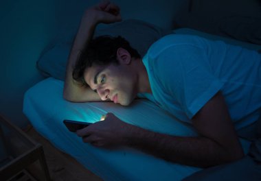 Addicted Man sohbet ve yatakta gece geç saatlere kadar akıllı telefon ile Internette sörf. Sıkılmış, uykusuz ve kasvetli ışık ile karanlık odada yorgun. Uykusuzluk ve mobil bağımlılık kavramı.