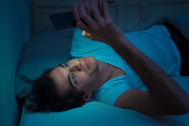 Sosyal medya genç adam sohbet ve yatakta gece akıllı telefon Internette sörf Addicted. Mobil ekran ışığı ile karanlık yatak odasında uykusuz. Uykusuzluk ve çevrimiçi ağ bağımlılığı.