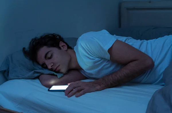 ベッドで夜にスマート携帯電話で眠りに落ちるソーシャルメディアの若者に中毒 モバイル画面のライトが点灯した暗い寝室で眠っている男のライフスタイルの肖像画 モバイル利用中毒 — ストック写真
