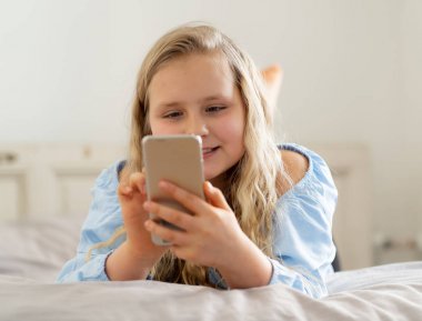 Cep telefonu oynarken güzel sarışın mutlu küçük kız. Sevimli çocuk evde yatakta yatan Smartphone sosyal medya uygulaması ile internette sohbet. Teknoloji eğitimi ve çocuk mobil istismar.