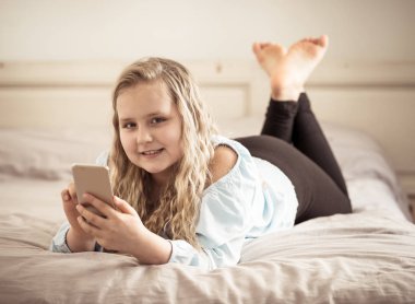 Cep telefonu oynarken güzel sarışın mutlu küçük kız. Sevimli çocuk evde yatakta yatan Smartphone sosyal medya uygulaması ile internette sohbet. Teknoloji eğitimi ve çocuk mobil istismar.