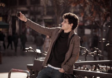 Heyecanlı Trendy genç adam selfie veya Avrupa şehir Caddesi veya üniversite kampüsünde tatil akıllı cep telefonu bloglama alarak. Tatil ve teknoloji bağlantı ve sosyal medya konsepti.