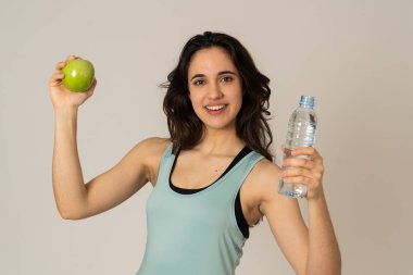 Mutlu fitness kadın güçlü ve sağlıklı duygu bir elma ve su şişesi tutarak gülümseyen. Sağlıklı yaşam tarzı, egzersiz ve beslenme kavramı. Nötr arka planda izole latin modeli portresi.