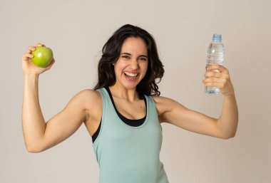 Mutlu fitness kadın güçlü ve sağlıklı duygu bir elma ve su şişesi tutarak gülümseyen. Sağlıklı yaşam tarzı, egzersiz ve beslenme kavramı. Nötr arka planda izole latin modeli portresi.
