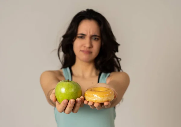 Beautiful young fit latin woman tempted having to make choice; apple and doughnut, healthy or unhealthy food. Fitness and nutrition healthy lifestyle and Diet concept. Studio shot with copy space.