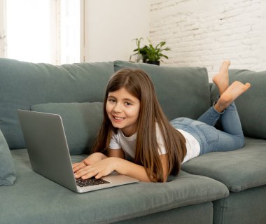 Güzel sevimli sevimli kız öğrenci oynarken ve dizüstü bilgisayar da internette sörf gülümseyerek ve evde kanepede yatarken. Dijital teknoloji, internet kullanımı ve yaşam tarzı konsepti.