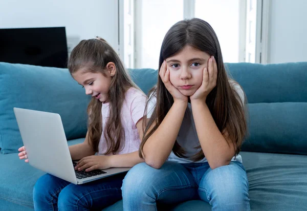 Sibling angry at younger sister spending too much time online using laptop. Digital technology addicted kid playing with computer ignoring her sad older sister. In children and internet addiction.