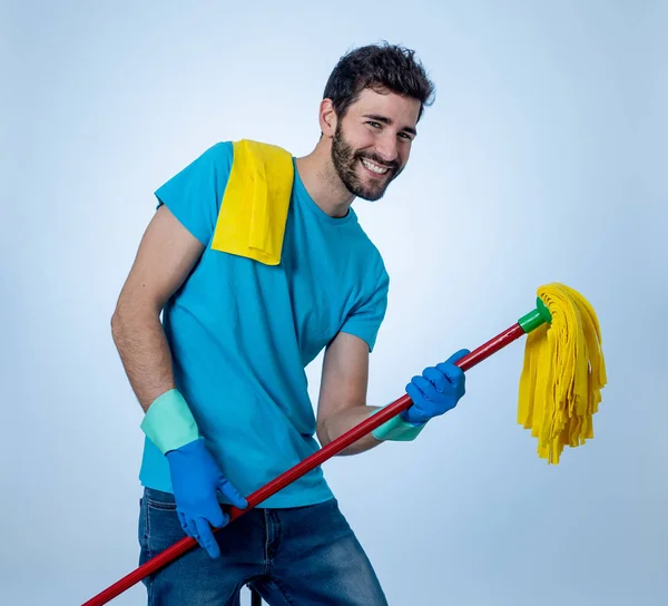 Breaking gender stereotypes. Portrait of happy young man having fun cleaning and doing housework holding a mop as microphone singing and dancing. In Changing men and womens roles in society.