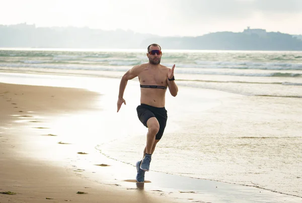 Athlete runner running with heart rate monitor and smart watch on beach. Shirtless fitness man exercising in outdoor training. Advertising style in sports technology healthcare wellness innovation.