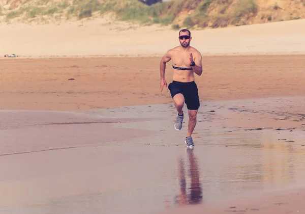 Athlete runner running with heart rate monitor and smart watch on beach. Shirtless fitness man exercising in outdoor training. Advertising style in sports technology healthcare wellness innovation.