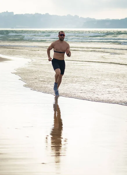 Sports fitness man with heart rate monitor running on beach with smart watch. Fit body male runner jogging by the seashore in workout exercise and training technology innovation in healthy lifestyle.