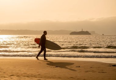 Gün batımında veya gün doğumunda deniz kıyısında sörf tahtası ile yürüyen sörfçü. Yüksek dalgalar arayan sörf adam silueti. Açık hava su sporları, tatil ve macera yaşam tarzı konsepti.