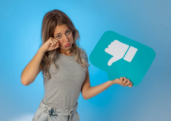 Attractive unhappy young woman holding social media symbol of dislike feeling sad and rejected in internet obsession, social network notification icons, cyber bullying and technology communication.