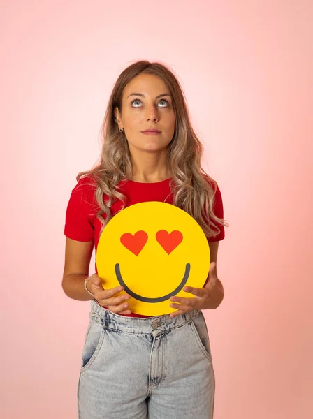 Attractive young woman holding a Hearts Eyes or In Love face emoji feeling happy and excited sharing it in social media. Facial expressions, social network notification icon and technology concept.