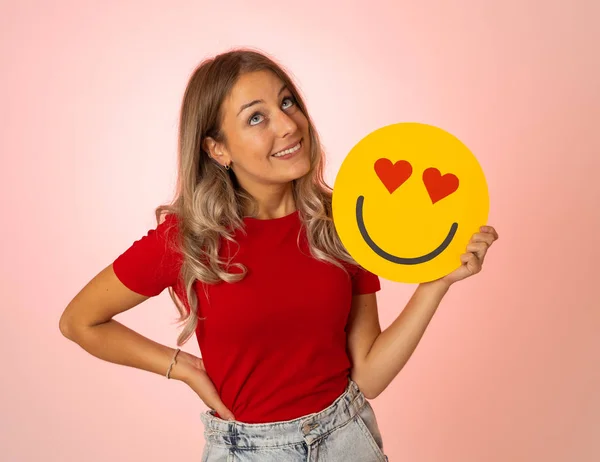 Attractive young woman holding a Hearts Eyes or In Love face emoji feeling happy and excited sharing it in social media. Facial expressions, social network notification icon and technology concept.