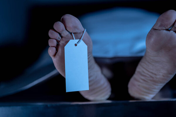 Close-up of dead body feet at morgue or hospital with toe label or information ring and identification blank tag. Cadaver lying on steal table covered with sheet on autopsy table. Death concept.