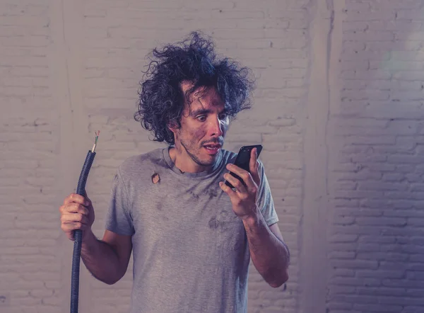 Portrait of young man holding electrical cable after domestic accident with dirty burnt funny face expression calling desperate with mobile phone asking for help. Electricity repairs and DIY concept.