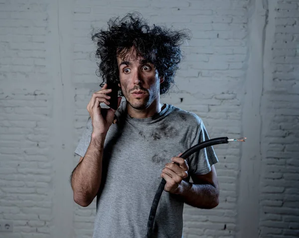 Portrait of young man holding electrical cable after domestic accident with dirty burnt funny face expression calling desperate with mobile phone asking for help. Electricity repairs and DIY concept.