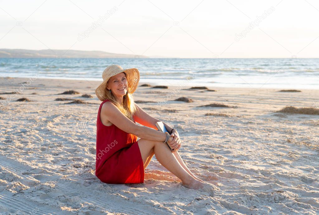 Happy attractive Mature woman in red dress enjoying outdoors and freedom on the beach, open arms outstretched in hope after coronavirus quarantine eased. Back to life, outdoors and new normal concept.