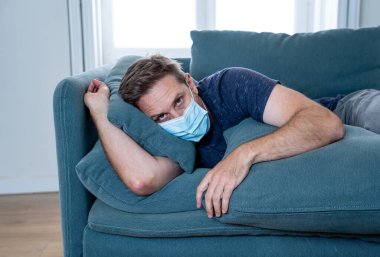 Oturma odasındaki kanepede koruyucu maskeli üzgün bir adam. Koronavirüs kilitlenmesi ve sosyal mesafeler arasında bunalıma girmiş yorgun ve endişeli hissediyor. Ruh sağlığı ve tecrit kavramı.