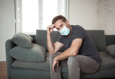Oturma odasındaki kanepede koruyucu maskeli üzgün bir adam. Koronavirüs kilitlenmesi ve sosyal mesafeler arasında bunalıma girmiş yorgun ve endişeli hissediyor. Ruh sağlığı ve tecrit kavramı.