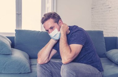 Oturma odasındaki kanepede koruyucu maskeli üzgün bir adam. Koronavirüs kilitlenmesi ve sosyal mesafeler arasında bunalıma girmiş yorgun ve endişeli hissediyor. Ruh sağlığı ve tecrit kavramı.