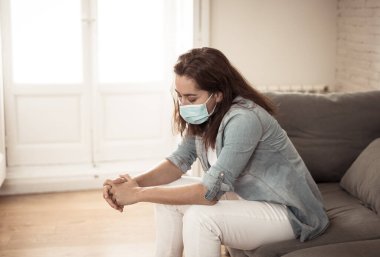 Oturma odasındaki kanepede koruyucu maskeli üzgün Latin kadın. Koronavirüs hapsi ve sosyal mesafeler yüzünden bunalımda ve yorgun hissediyor. Ruh sağlığı ve tecrit kavramı.