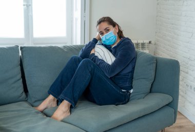 Oturma odasındaki kanepede koruyucu maskeli üzgün Latin kadın. Koronavirüs hapsi ve sosyal mesafeler yüzünden bunalımda ve yorgun hissediyor. Ruh sağlığı ve tecrit kavramı.