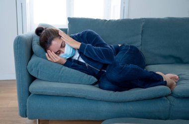 Oturma odasındaki kanepede koruyucu maskeli üzgün Latin kadın. Koronavirüs hapsi ve sosyal mesafeler yüzünden bunalımda ve yorgun hissediyor. Ruh sağlığı ve tecrit kavramı.