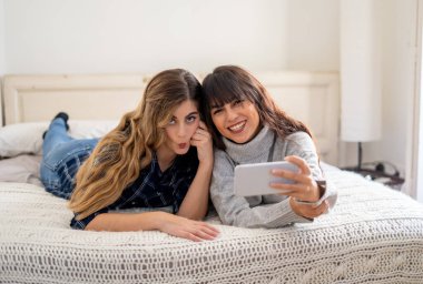 COVID-19, evde kal. Bağlantıda kal. Mutlu kadın kız arkadaşlar Coronavirus kilitleme ve sosyal mesafeler sırasında cep telefonu kullanarak arkadaşlarıyla görüntülü konuşma ya da çevrimiçi grup ev partileri yapıyorlar..