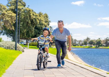 Oğlan babasıyla gölün yanındaki parkta bisiklet sürmeyi öğreniyor. Baba ve oğul bisiklette birlikte eğleniyorlar. Mutlu aile, açık hava aktiviteleri, çocukluk ve ebeveynlik kavramı.