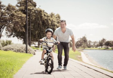 Oğlan babasıyla gölün yanındaki parkta bisiklet sürmeyi öğreniyor. Baba ve oğul bisiklette birlikte eğleniyorlar. Mutlu aile, açık hava aktiviteleri, çocukluk ve ebeveynlik kavramı.