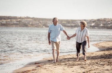 Gün batımında boş kumsalda el ele tutuşan ve el ele tutuşan mutlu yaşlı çift. Aktif, sağlıklı yaşlı kadın ve erkek deniz kenarında romantik bir yürüyüş yapıyorlar. Birlikte yaşlanma ve emeklilik yaşam tarzı konsepti.