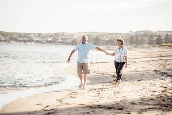 快乐的老夫妇在日落时拥抱并牵着手在空旷的海滩上散步 积极健康的老年男女在海上浪漫地散步 共同老龄化和退休生活方式概念 — 图库照片