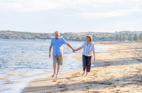 快乐的老夫妇在日落时拥抱并牵着手在空旷的海滩上散步 积极健康的老年男女在海上浪漫地散步 共同老龄化和退休生活方式概念 — 图库照片