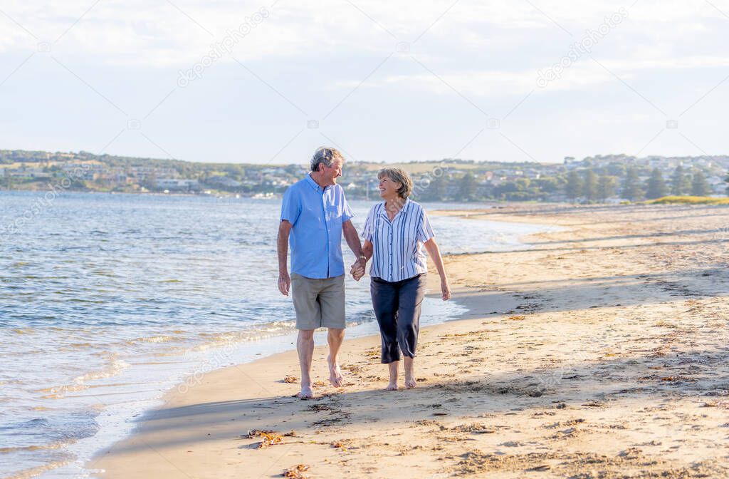 Happy senior couple embracing and holding hands walking on empty beach at sunset. Active healthy elderly woman and man on a romantic walk by the sea. Aging together and retirement lifestyle concept.