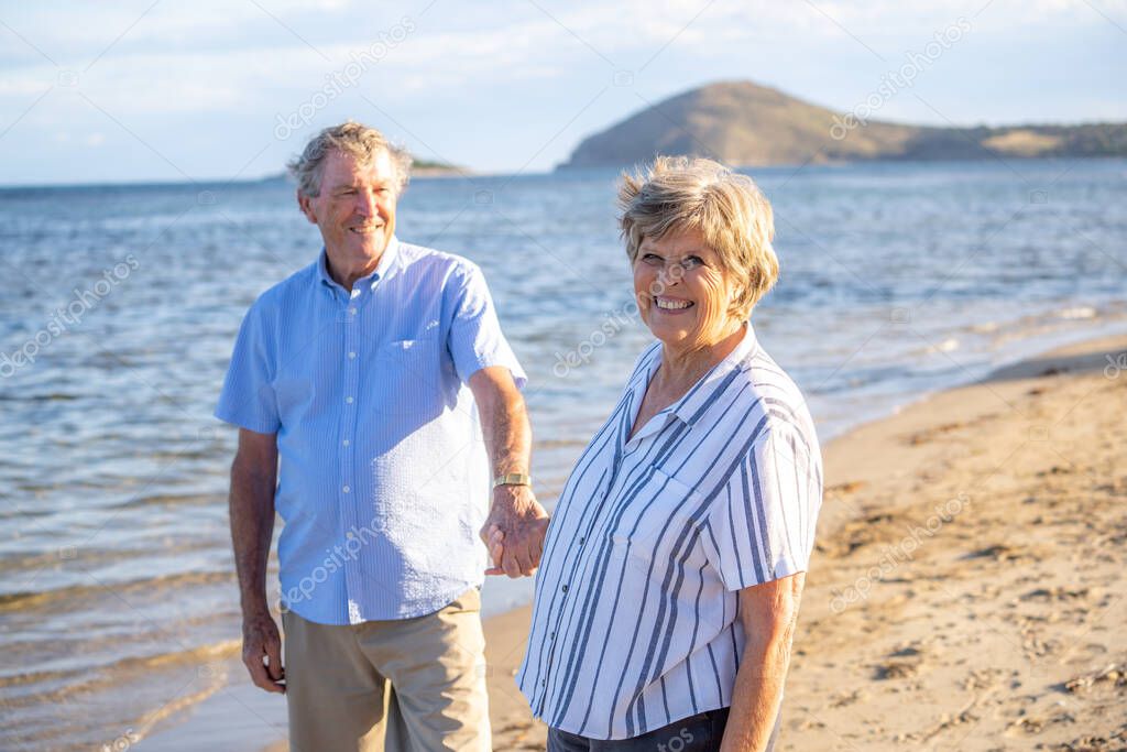 Happy senior couple embracing and holding hands walking on empty beach at sunset. Active healthy elderly woman and man on a romantic walk by the sea. Aging together and retirement lifestyle concept.