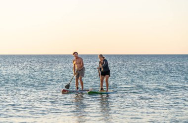 SUP kürekli sörf tahtasında, gün batımında sakin denizde güzel kumsalda eğlenen olgun beyaz bir çift. Aktif modern yaşlı insanlar, açık hava aktiviteleri, birlikte yaşlanma ve sağlıklı yaşam tarzı..