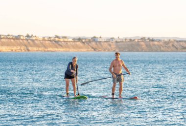 SUP kürekli sörf tahtasında, gün batımında sakin denizde güzel kumsalda eğlenen olgun beyaz bir çift. Aktif modern yaşlı insanlar, açık hava aktiviteleri, birlikte yaşlanma ve sağlıklı yaşam tarzı..