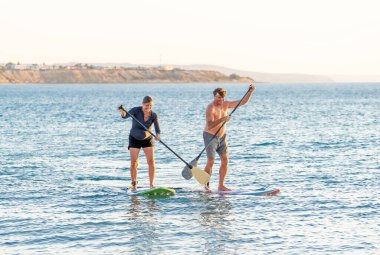 SUP kürekli sörf tahtasında, gün batımında sakin denizde güzel kumsalda eğlenen olgun beyaz bir çift. Aktif modern yaşlı insanlar, açık hava aktiviteleri, birlikte yaşlanma ve sağlıklı yaşam tarzı..