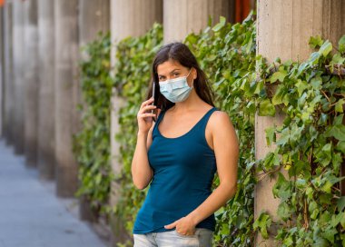 COVID-19 salgını. Şehir sokaklarında koruyucu yüz maskesi takarak yürüyen ve şehir yazında cep telefonu kullanan genç bir kadın. Coronavirus, Yeni Normal ve zorunlu yüz maskesi kullanımı