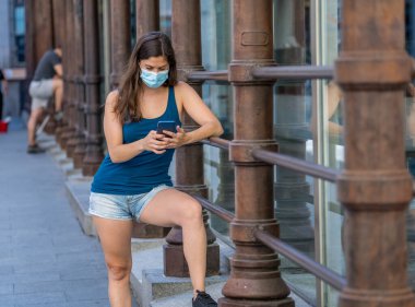 COVID-19 salgını. Şehir sokaklarında koruyucu yüz maskesi takarak yürüyen ve şehir yazında cep telefonu kullanan genç bir kadın. Coronavirus, Yeni Normal ve zorunlu yüz maskesi kullanımı