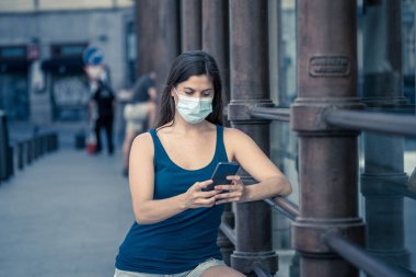 COVID-19 salgını. Şehir sokaklarında koruyucu yüz maskesi takarak yürüyen ve şehir yazında cep telefonu kullanan genç bir kadın. Coronavirus, Yeni Normal ve zorunlu yüz maskesi kullanımı