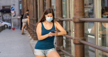 COVID-19 salgını. Şehir sokaklarında koruyucu yüz maskesi takarak yürüyen ve şehir yazında cep telefonu kullanan genç bir kadın. Coronavirus, Yeni Normal ve zorunlu yüz maskesi kullanımı