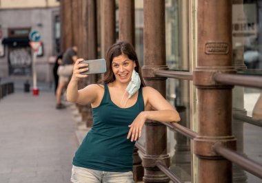 COVID-19 salgını. Şehir sokaklarında koruyucu yüz maskesi takarak yürüyen ve şehir yazında cep telefonu kullanan genç bir kadın. Coronavirus, Yeni Normal ve zorunlu yüz maskesi kullanımı