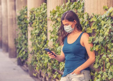 COVID-19 salgını. Şehir sokaklarında koruyucu yüz maskesi takarak yürüyen ve şehir yazında cep telefonu kullanan genç bir kadın. Coronavirus, Yeni Normal ve zorunlu yüz maskesi kullanımı